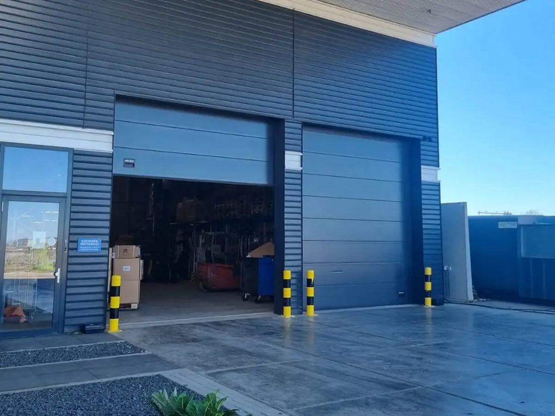 Blauw industrieel pakhuis met twee grote roldeuren en gele-zwarte gestreepte veiligheidsbolder(s).